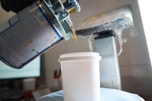 Male in protective gloves standing in painting booth, holding container of paint and pouring paint into bucket